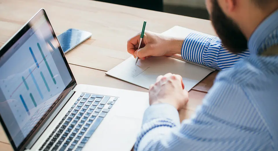 Nahaufnahme Mann in Hemd, der sich neben seinem Laptop auf einem Blatt Papier Notizen macht