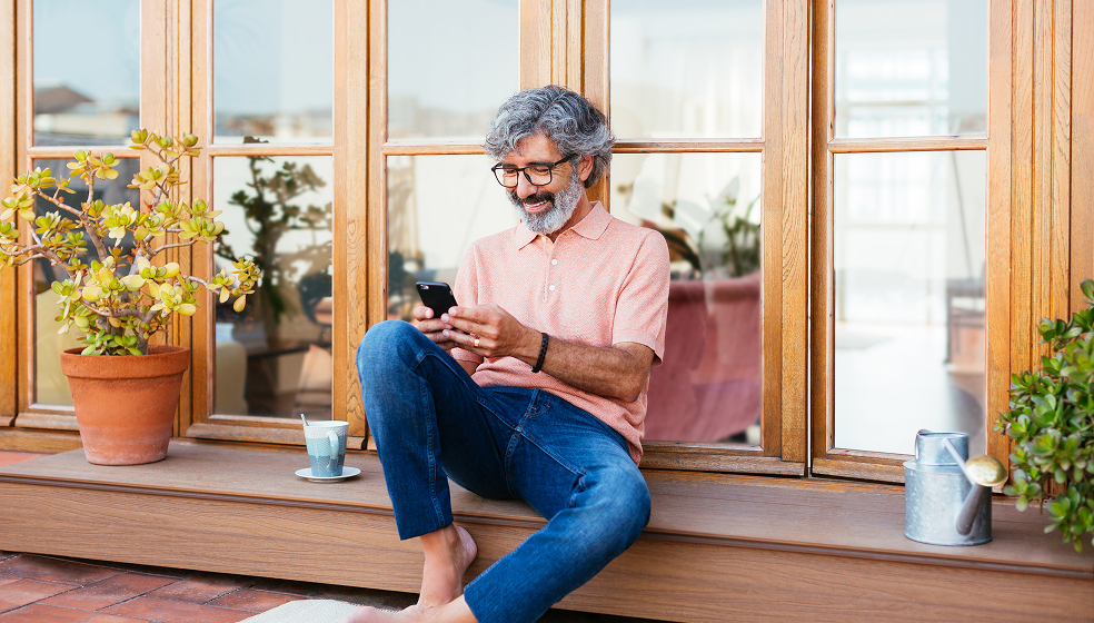 Mann in rosanem Hemd sitzt draußen vor seinem Terrassenfenster und schaut in sein Smartphone