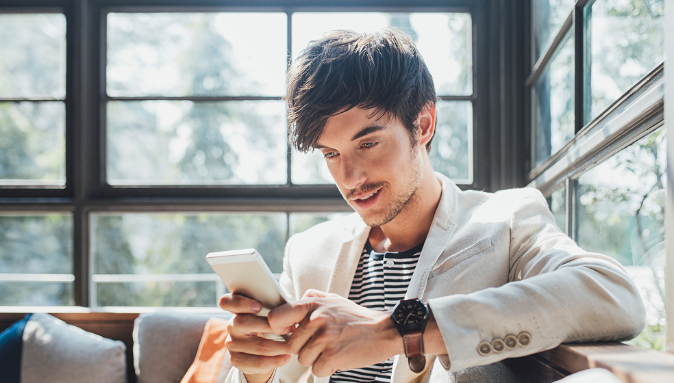Dunkelhaariger Mann in weißem Sakko sitzt auf einem Sofa und schaut in sein Smartphone