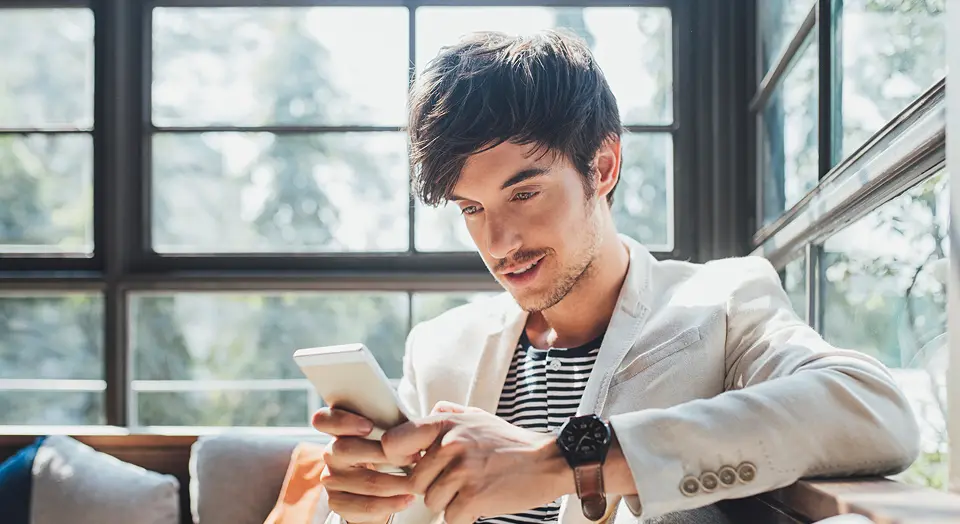 Dunkelhaariger Mann in weißem Sakko sitzt auf einem Sofa und schaut in sein Smartphone