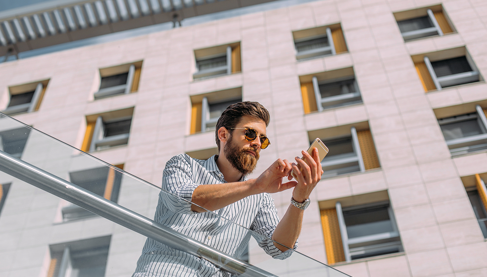 Mann in Hemd mit Sonnenbrille steht draußen an einem Geländer und schaut auf sein Smartphone