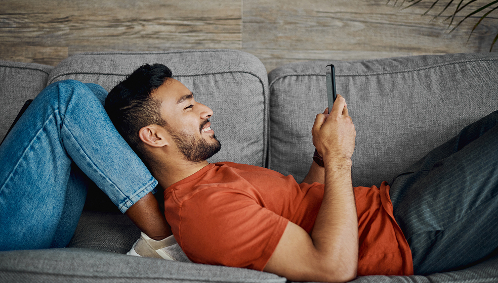 Mann in orangenem T-Shirt liegt auf einem Sofa und schaut in sein Smartphone