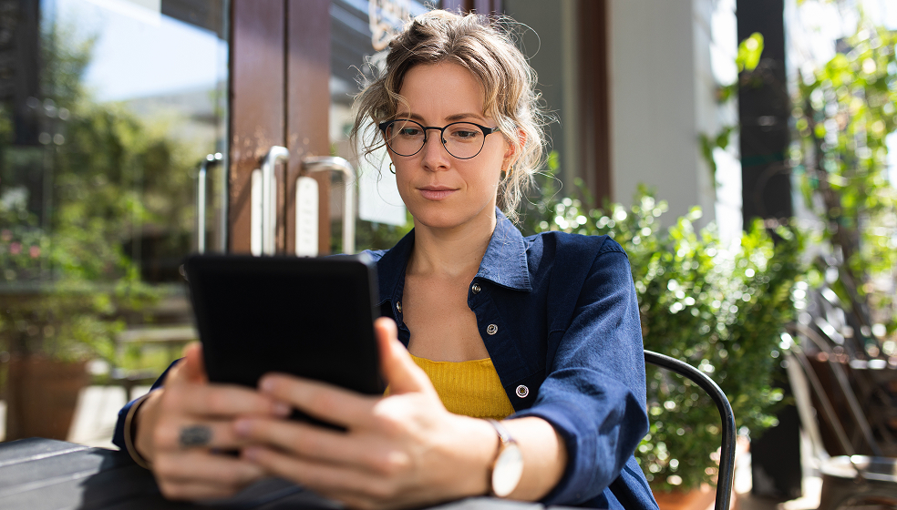 Frau mit Brille sitzt in einem grünen Café und schaut in ihr Tablet