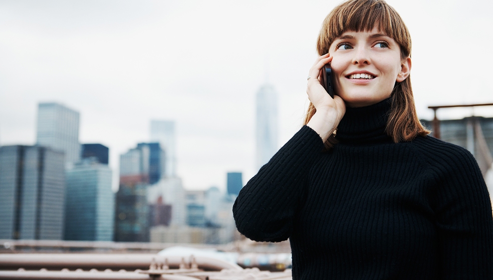 Frau in schwarzem Rollkragenpullover telefoniert im Freien vor einer großen City Skyline