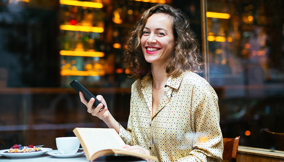 Lockige Frau in Hemd sitzt mit Notizbuch in einem Café und lächelt in die Kamera