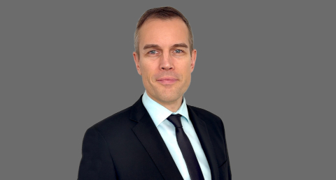 Ein Mann mit braunen Haaren den man Oberkörper aufwärts abgebildet sieht, trägt einen dunkelblauen Anzug mit dunkler Krawatte und lächelt in die Kamera 