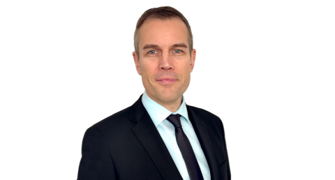 Ein Mann mit braunen Haaren den man Oberkörper aufwärts abgebildet sieht, trägt einen dunkelblauen Anzug mit dunkler Krawatte und lächelt in die Kamera 