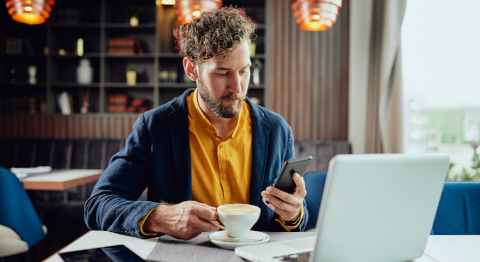 Das Bild zeigt eine Person, die an einem Tisch in einem modernen Café oder Restaurant sitzt. Die Person arbeitet an einem Laptop, hält ein Smartphone in einer Hand und eine Tasse Kaffee in der anderen. Im Hintergrund sind Bücherregale und hängende Lampen zu sehen. Die Atmosphäre ist hell und entspannt.