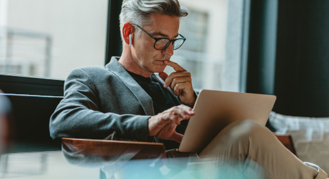 Grauhaariger Mann mit Brille sitzt in einem Sessel und arbeitet konzentriert an einem Laptop.