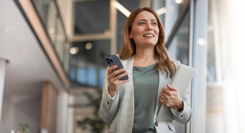 Frau mit hellbraunem Haar, hellgrauem Blazer und graugrünem T-Shirt hält in der einen Hand ein blaues Smartphone und in der anderen ein zugeklapptes, silbernes Notebook. Die Frau blickt mit einem Lächeln an der Kamera vorbei in die Ferne. Der Hintergrund zeigt einen hellen, modernen Innenraum.