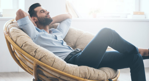 Ein Mann sitzt entspannt mit verschränkten Armen in einem großen, runden Korbsessel. Er trägt eine helle Bluse und dunkle Hose. Im Hintergrund ist ein helles Zimmer mit einem Fenster und einigen Gegenständen auf der Fensterbank zu sehen. Die Atmosphäre wirkt ruhig und gemütlich.