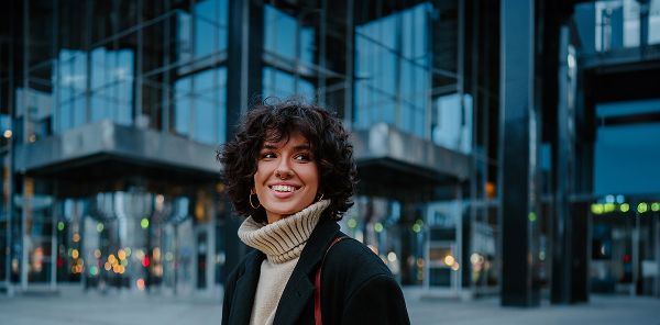 Das Bild zeigt eine Person mit kurzen, lockigen Haaren, die einen hellen Rollkragenpullover und einen dunklen Mantel trägt. Im Hintergrund ist eine moderne Glas- und Stahlarchitektur in einer städtischen Umgebung zu sehen, mit verschwommenen Lichtern und Reflexionen.