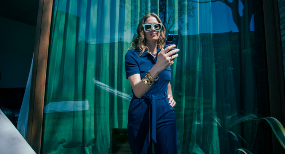 Frau mit langen lockigen rotblonden Haaren im blauen Kleid und auffälliger blauer Sonnenbrille geht an einem Fenster vorbei und blickt auf ihr Smartphone
