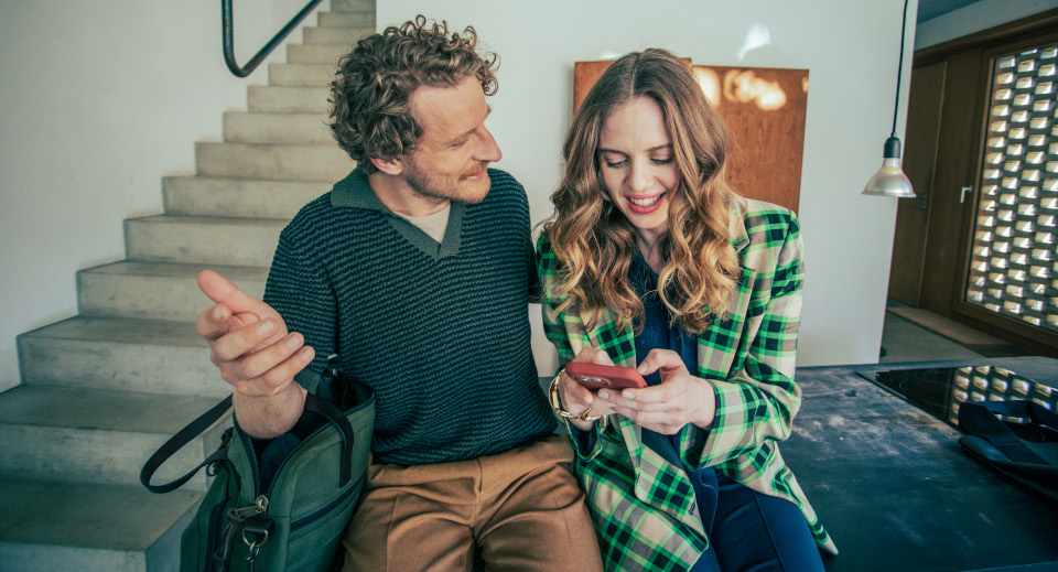 Junge Frau lächelt in die Kamera. Sie sitzt auf einer Auto Rückbank und durch das Fenster der Heckscheibe leuchtet der Sonnenuntergang. 