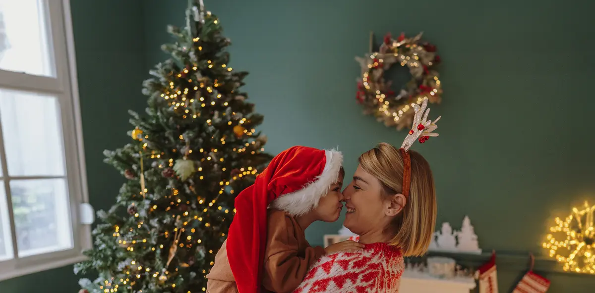 Mutter und Kind mit Weihnachtsmannmütze stupsen ihre Nasen aneinander im Hintergrund steht ein geschmückter Weihnachtsbaum und ein Kranz hängt an der Wandsbett, ein Mann und eine Frau mit bunter Verkleidung als Clown führen ihre Tricks auf. Der Mann pustet eine große Seifenblase, die Frau zeigt auf die Seifenblase..