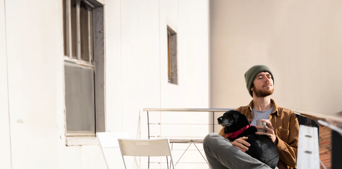 Mann mit Mütze sitzt mit seinem Hund auf dem Schoß auf dem Balkon und genießt das Wetter