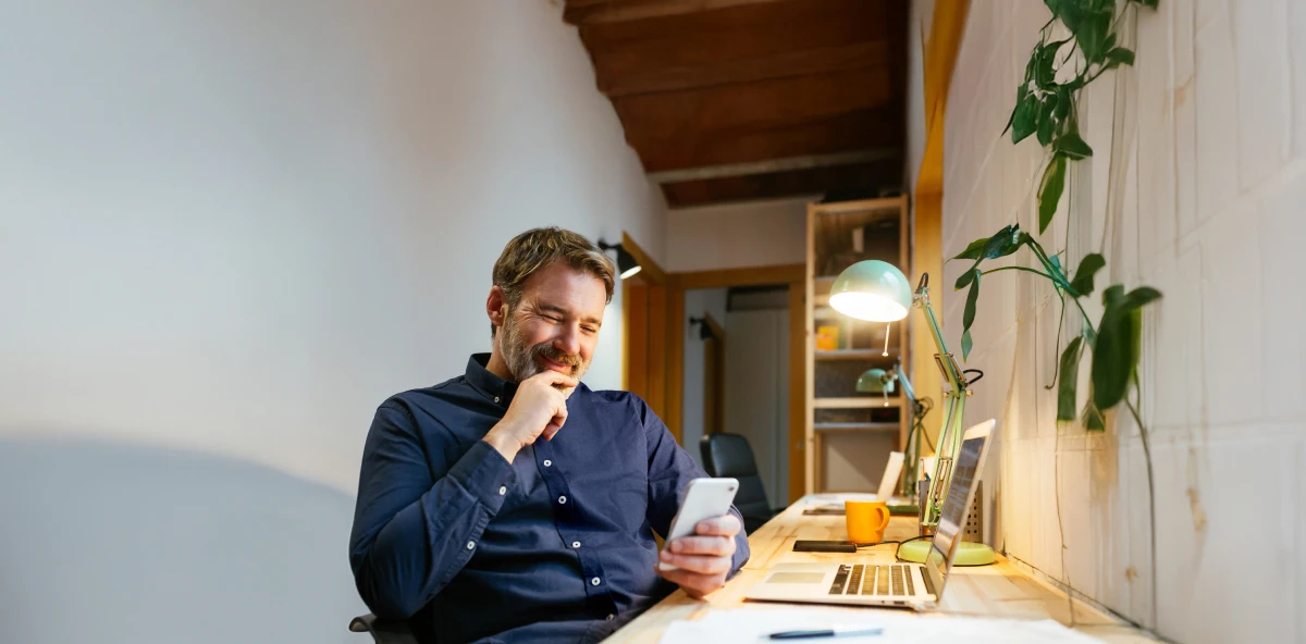 Mann in dunkelblauem Hemd sitzt am Schreibtisch und schaut in sein Smartphone