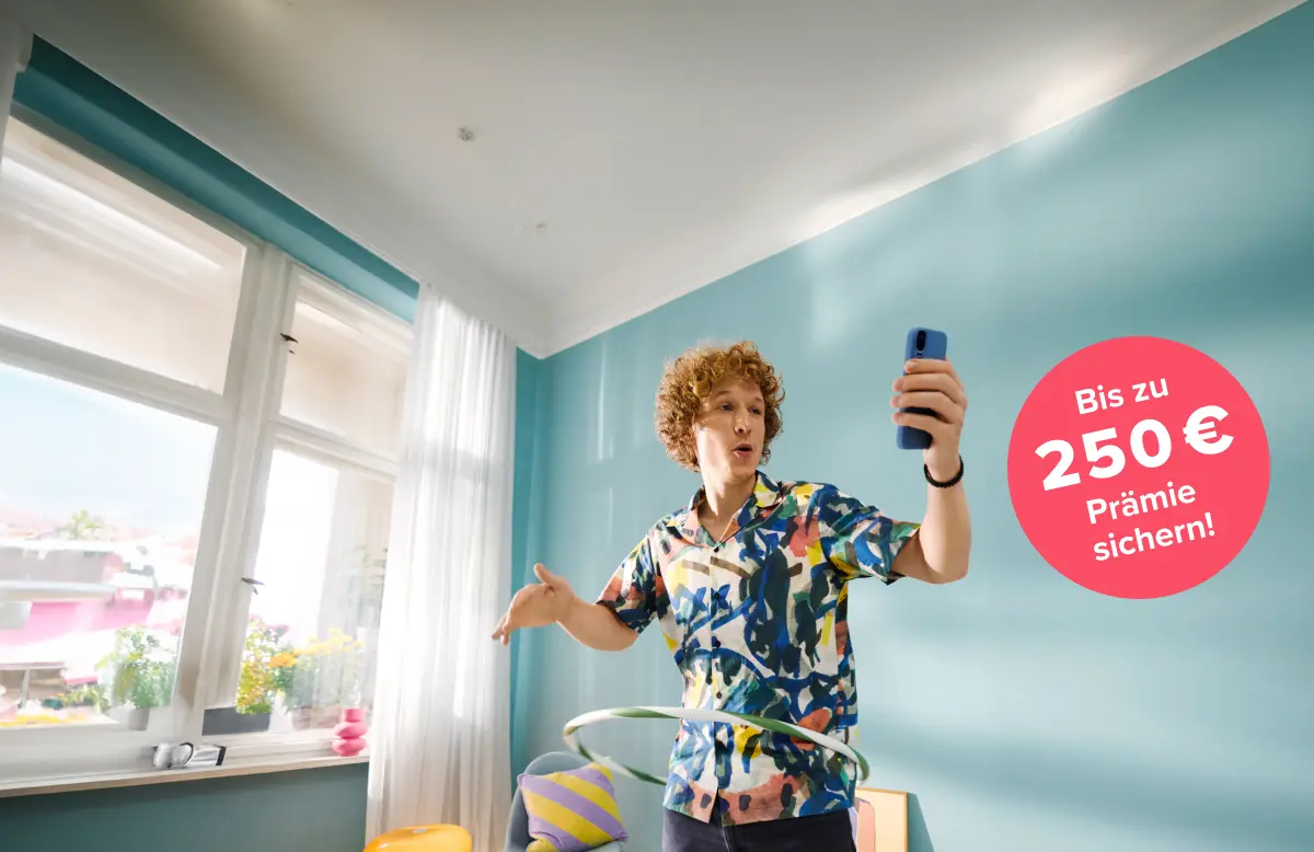 Mann mit orangenen Locken in buntem Hemd steht auf dem Sofa und schaut in sein Smartphone