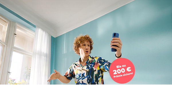 Mann mit orangenen Locken in buntem Hemd steht auf dem Sofa und schaut in sein Smartphone