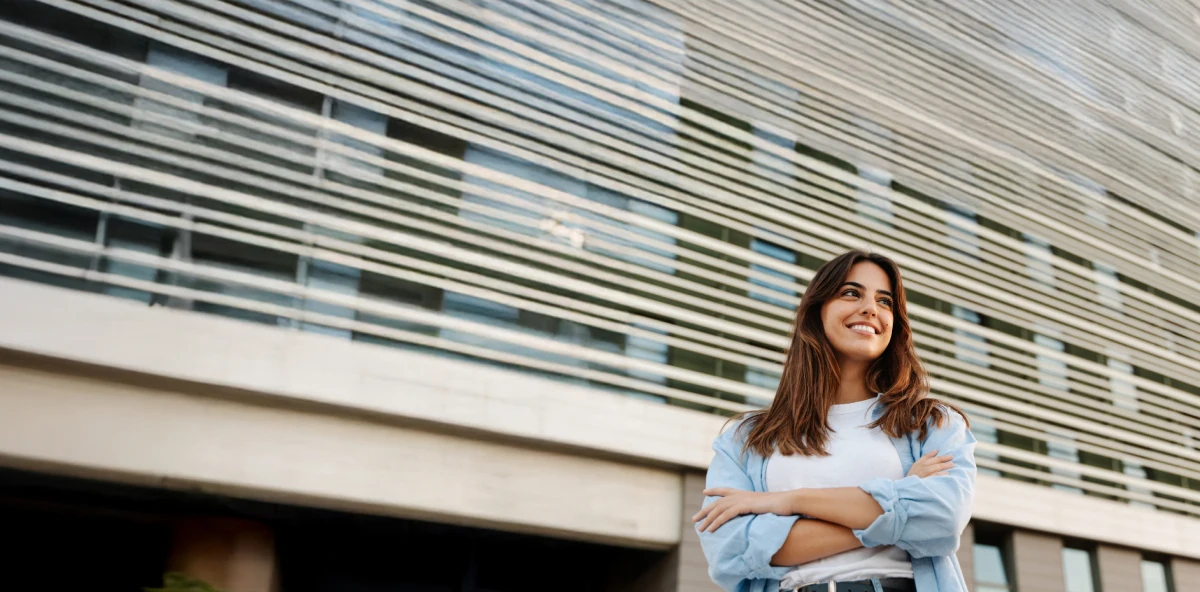 Braunhaarige Frau steht mit einem Lächeln und verschränkten Armen vor einem Gebäude und schaut in die Ferne