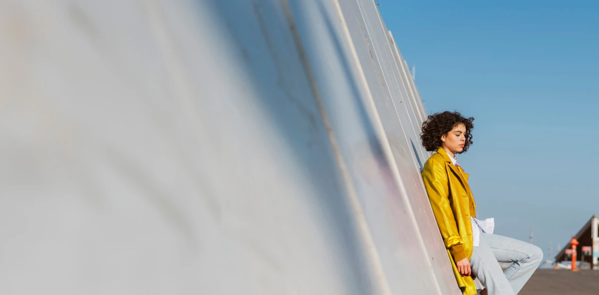 Frau in gelbem Mantel steht draußen angelehnt an eine schräge Wand