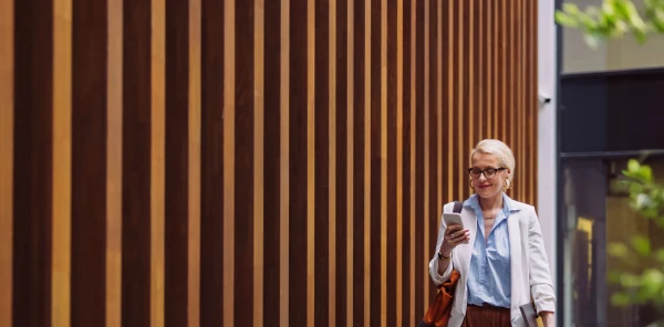 Frau in Businessklamotten läuft draußen in der Sonne vor einer Holzwand mit Smartphone in der Hand