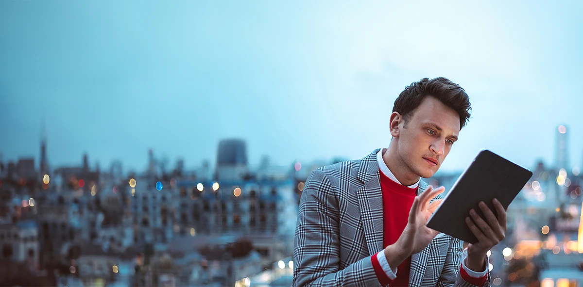 Mann in kariertem Sakko und roten Pullover tippt vor einer City-Skyline etwas in sein Tablet ein