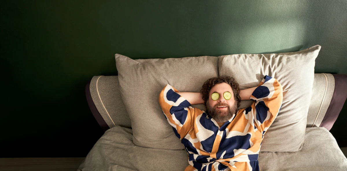 Lockiger Mann in buntem Hemd liegt entspannt mit Armen hinter dem Kopf und Gurkenscheiben auf den Augen auf seinem Bett