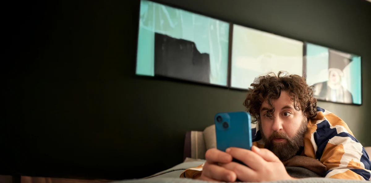 Mann mit Locken in buntem Hemd liegt auf dem Bauch auf dem Bett und schaut in sein blaues Smartphone