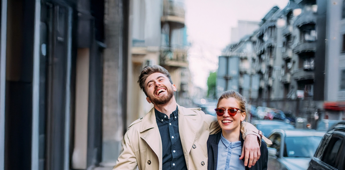 Mann mit kurzen braunen Haaren legt Arm um Frau mit Sonnenbrille und beide laufen lachend auf der Straße