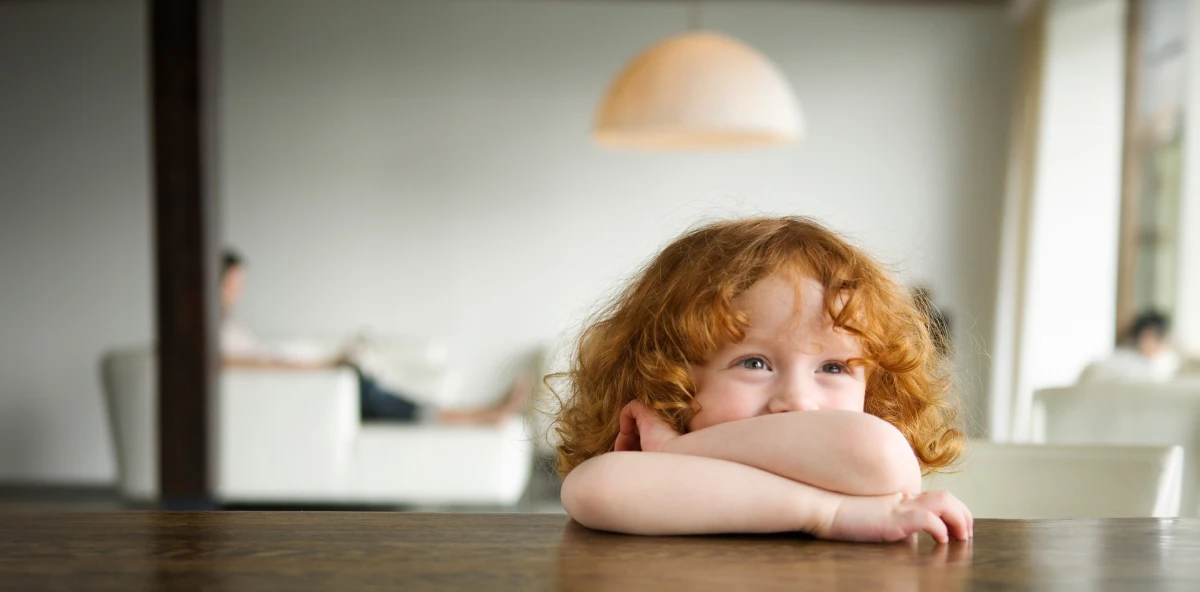 Kind mit orangenen Locken liegt mit den Armen abgestützt auf einem Tisch und lacht