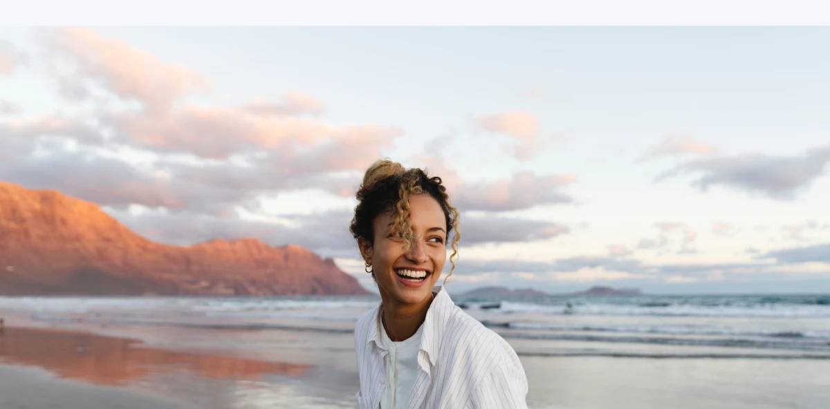 Frau mit lockigen Haaren lacht bei Sonnenuntergang an einem Strand