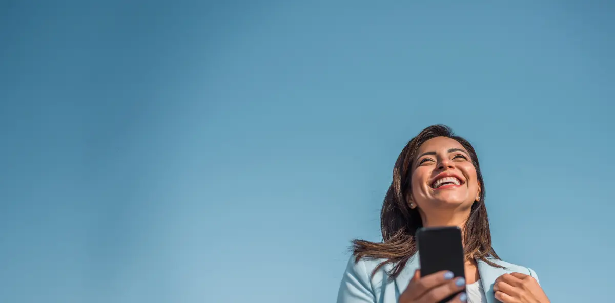 Braunhaarige Frau in Blazer von unten fotografiert hält Smartphone in der Hand und lächelt