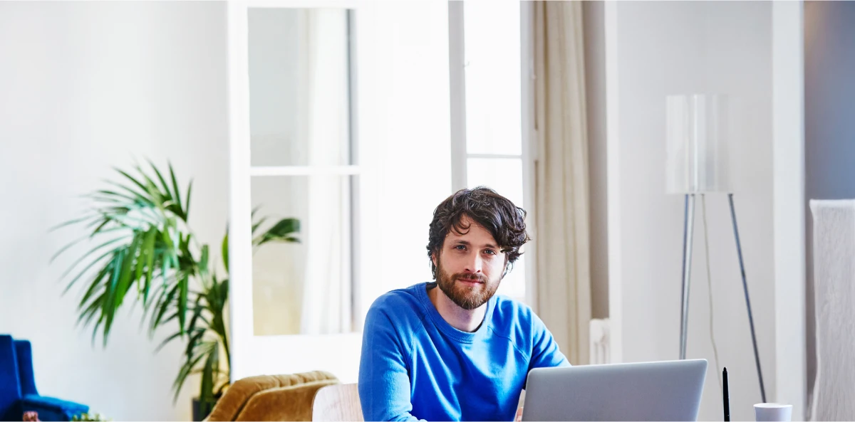Braunhaariger Mann in blauem Pullover sitzt in seinem Wohnzimmer am Laptop und schaut in die Kamera
