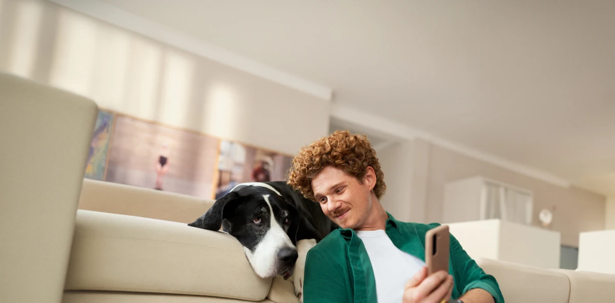 Mann in grünem Hemd mit orangenen Locken sitzt vor dem Sofa angelehnt an einen schwarz-weißen Hund