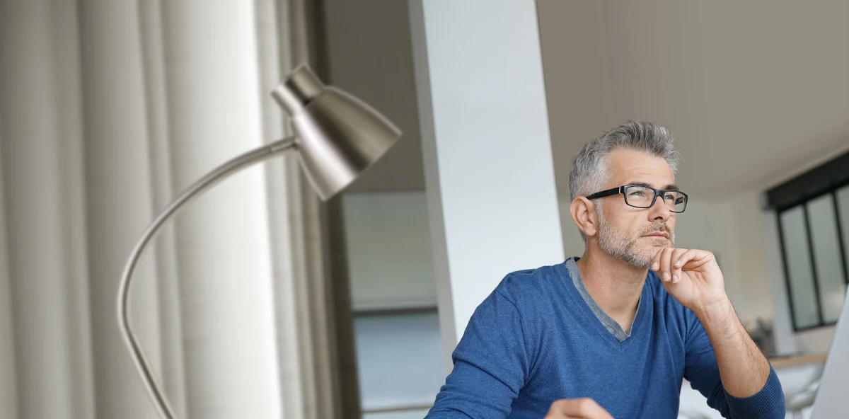 Grauhaariger Mann in blauem Pullover mit schwarzer Brille sitzt an seinem Laptop und schaut in die Ferne
