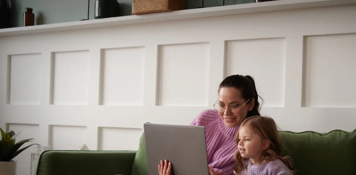 Frau mit dunklen Haaren und Brille sitzt mit einem Kind auf einem grünen Sofa und beide schauen auf einen Laptopbildschirm
