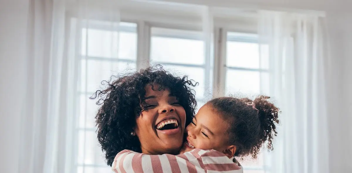 Frau mit dunklen Haaren wird von Kind mit dunklen Haaren umarmt und beide lachen, im Hintergrund ein Fenster