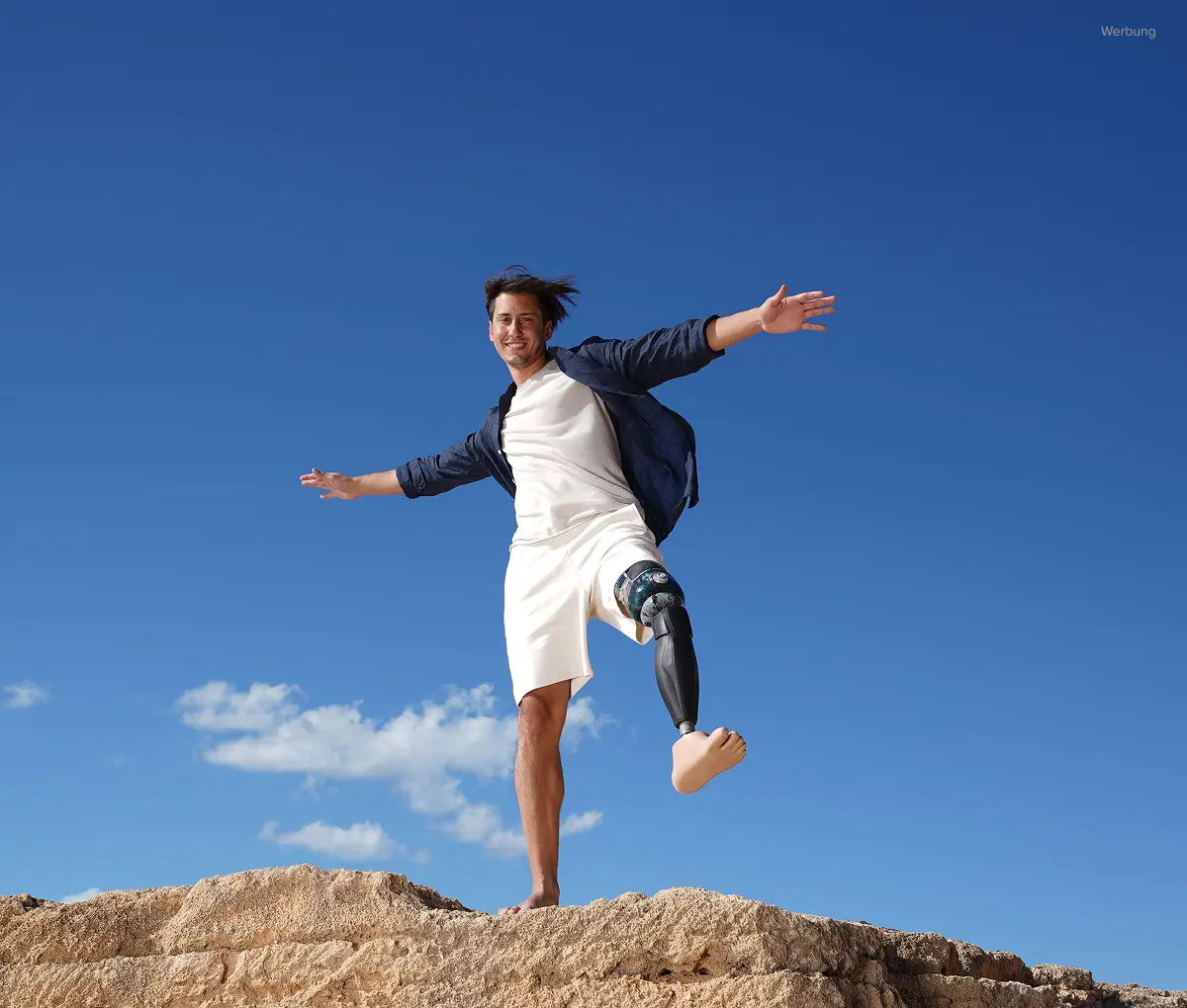 Junger Mann mit braunen Haaren und Beinprothese steht mit ausgebreiteten Armen vor blauem Himmel mit weißen Wolken