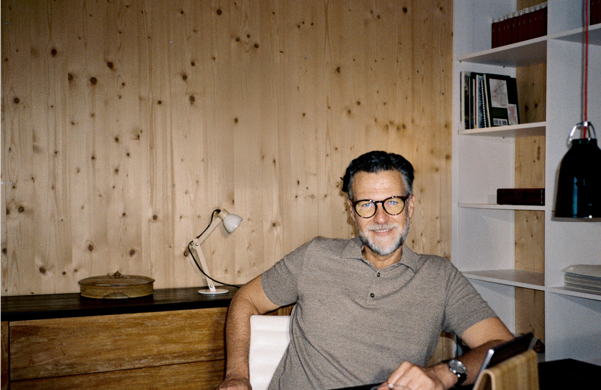 Mann mit Brille sitzt hinter einem Tisch und schaut lächelnd in die Kamera, im Hintergrund ist eine weißes Regal