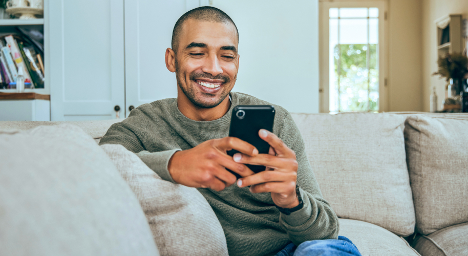 Mann sitzt lächelnd auf einem Sofa und schaut in sein Smartphone