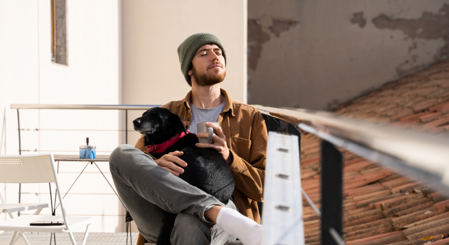 Mann mit Mütze sitzt mit Hund auf dem Schoß auf dem Balkon und genießt die Sonne